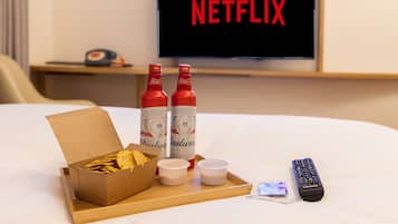 A hotel room with a bed, a tray with snacks, two Budweiser bottles, two small cups, a remote control, and a TV displaying the Netflix logo.