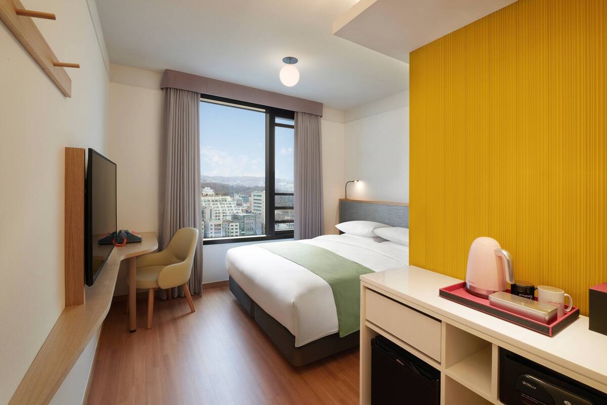 Quarto de hotel com cama, escrivaninha, cadeira, televisão e janela com vista para a cidade.