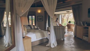 A bedroom with a bed, bedside table, lamp, and a view of the outdoors. - Lilayi Lodge (Lusaka)
