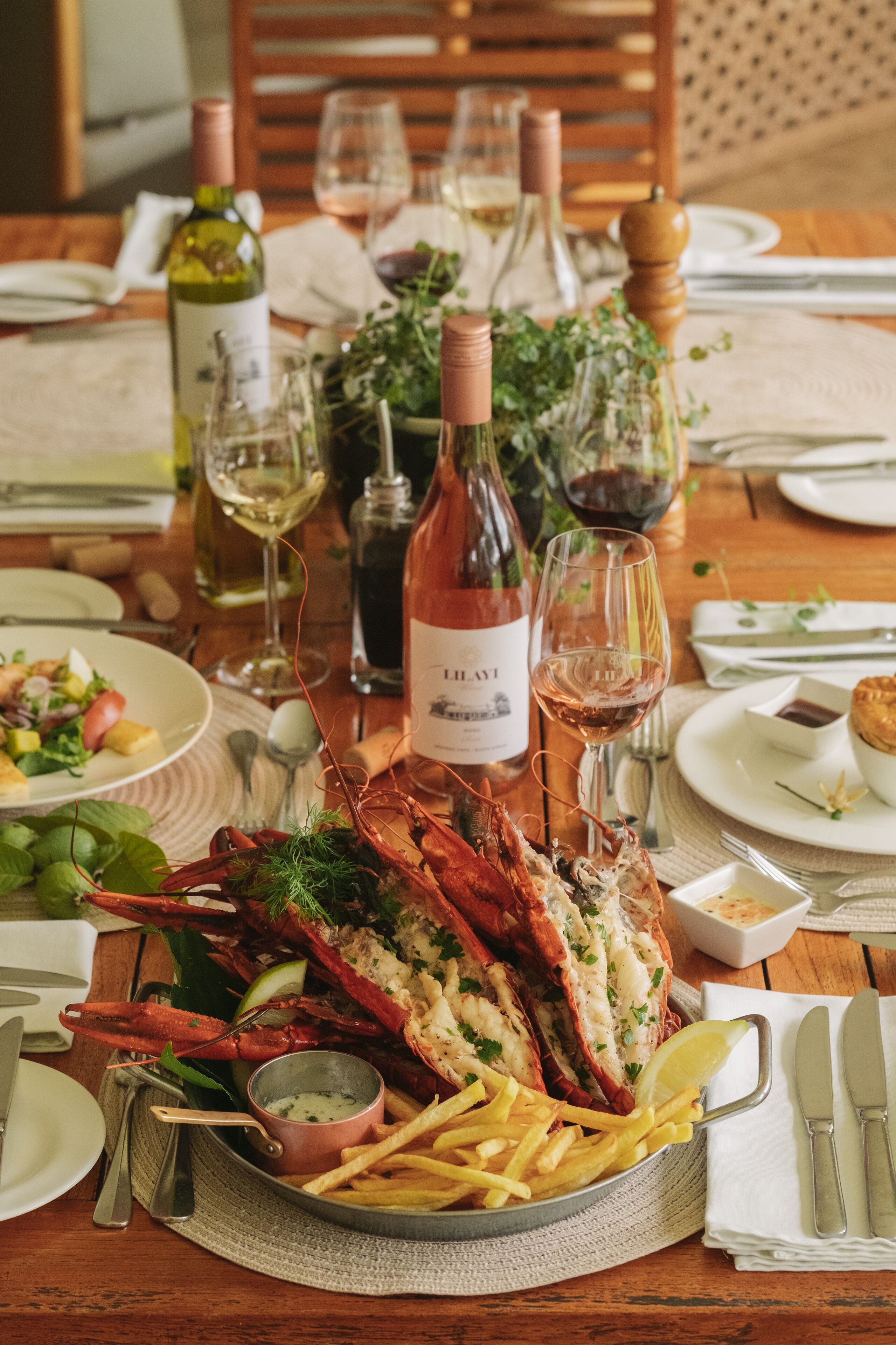 A seafood platter with lobster, lemon, and fries, accompanied by wine glasses and bottles on a wooden table.