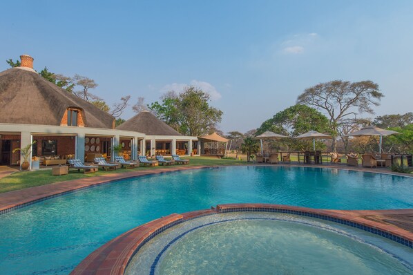 A swimming pool with lounge chairs, a thatched-roof building, and a clear sky. - Lilayi Lodge (Lusaka)