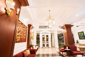Un vestíbulo de hotel con una imponente araña, sofás tapizados en rojo y un espejo grande con diseño de pan de oro.