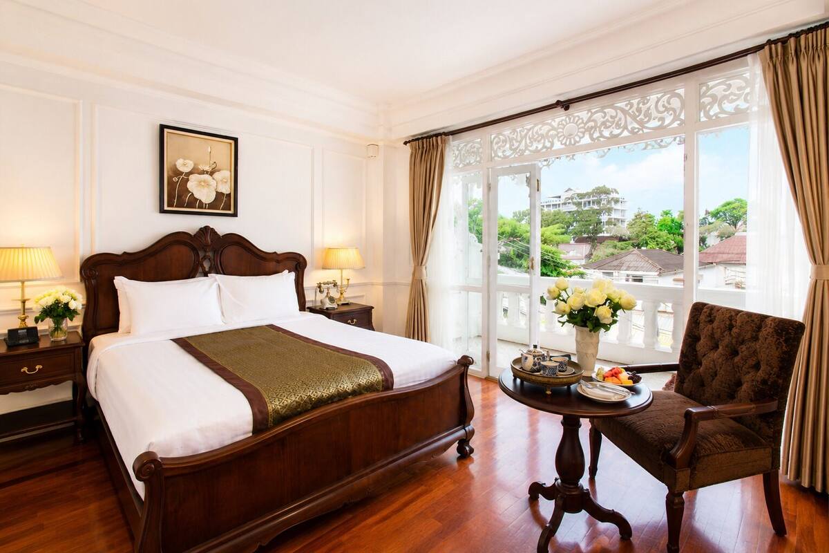 Hotelkamer met een groot bed, een zithoek met een fauteuil, een tafel met een bloevas, en uitzicht op een gebouw door het raam.