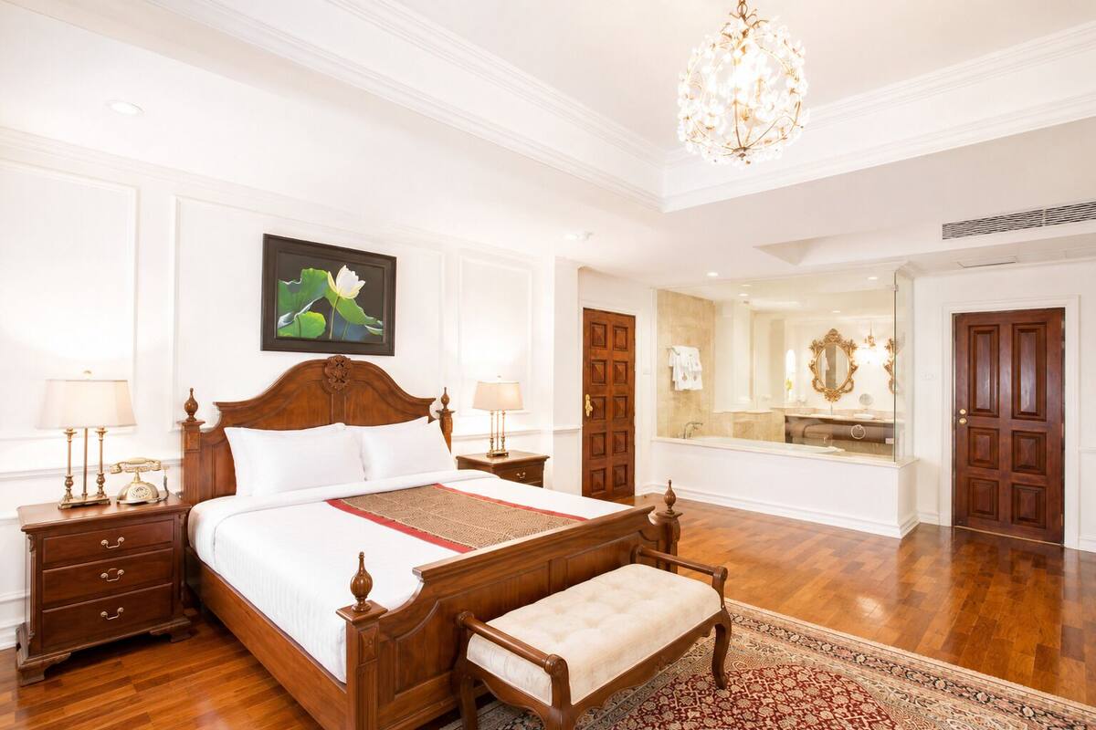 Een ruime slaapkamer met een groot bed, houten nachtkastjes, een bankje, een kroonluchter en een badkamer met spiegel en wastafel.
