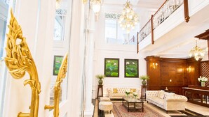 Um hall imponente com teto alto, lustres, uma escadaria monumental e uma área de estar com uma mesa de centro e sofás.