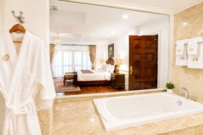 Habitación de hotel con un espejo grande, una cama, una mesita de noche, una lámpara, una puerta de madera y un baño con bañera.