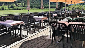 An outdoor seating area with tables and chairs, umbrellas, and a view of a mountainous landscape.