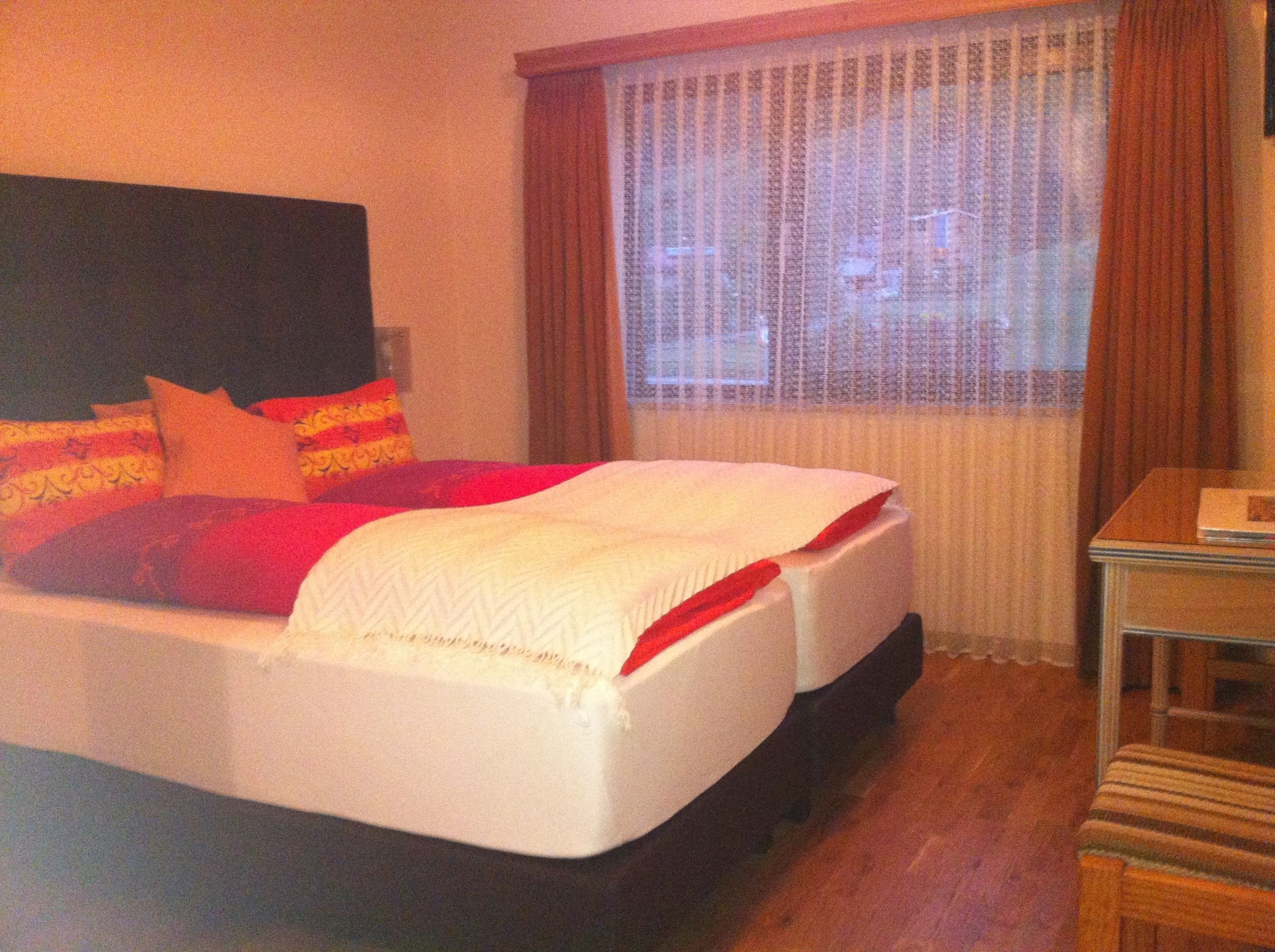 Cama bem arrumada com edredom vermelho e branco, cabeceira de madeira e janela com cortinas.