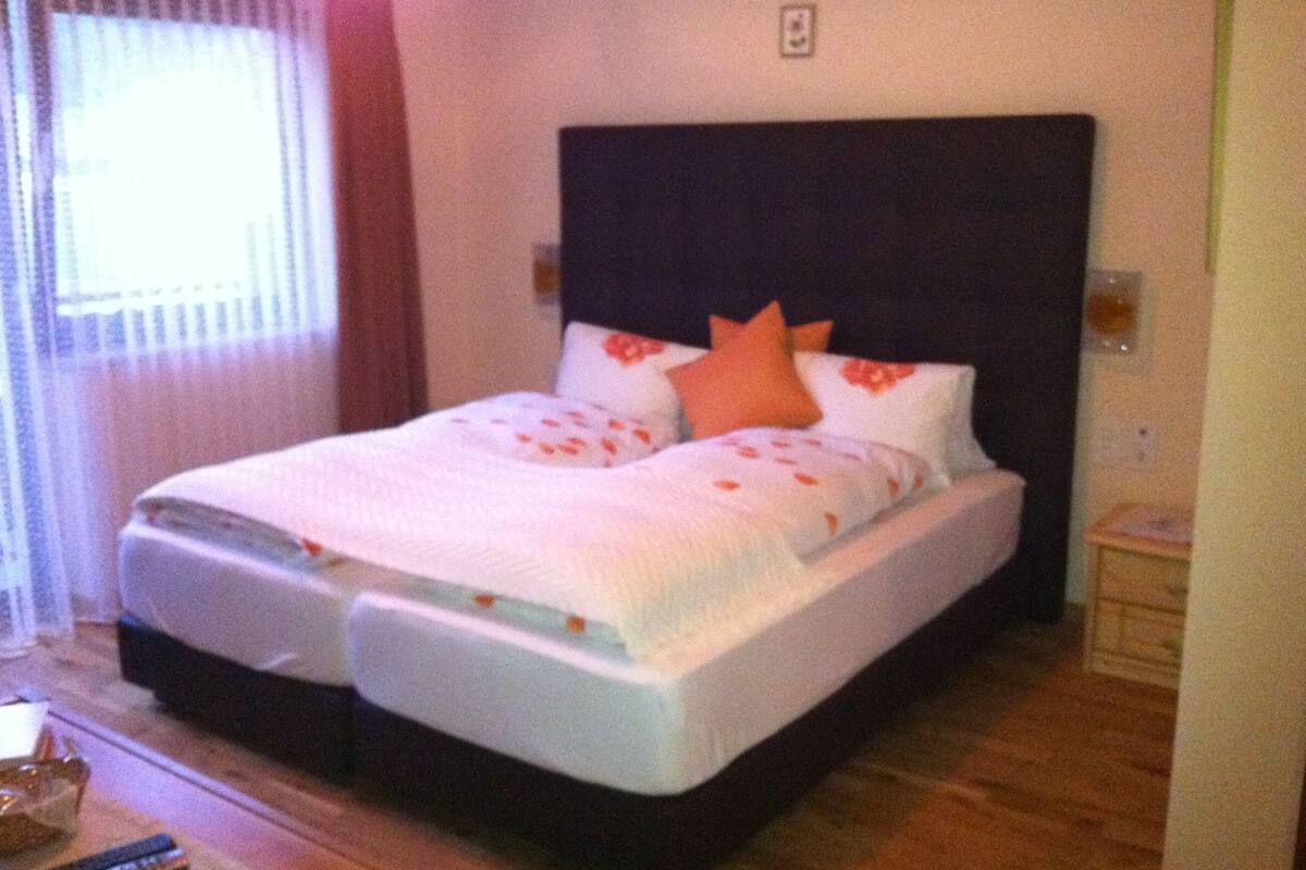 Ein Doppelbett mit dunkelbraunem Kopfteil, weißer Bettwäsche und orangefarbenen Kissen. Ein Nachttisch mit Lampe steht daneben.
