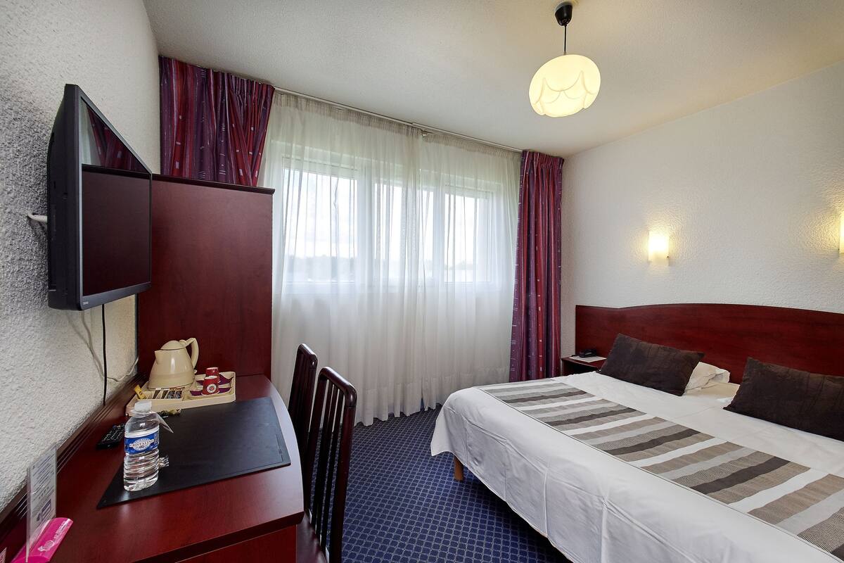 A hotel room with a bed, a desk with a chair, a television, and a window with curtains.