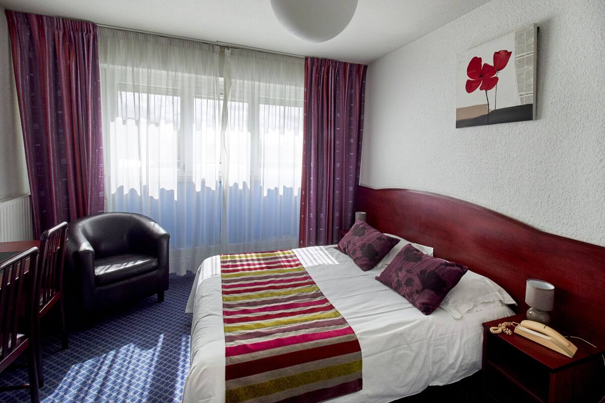A hotel room with a bed, a chair, a bedside table with a lamp, and a painting on the wall.