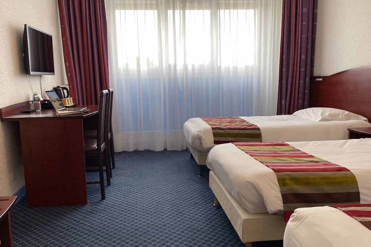 A hotel room with two beds, a desk with a laptop, a television, and a window with curtains.