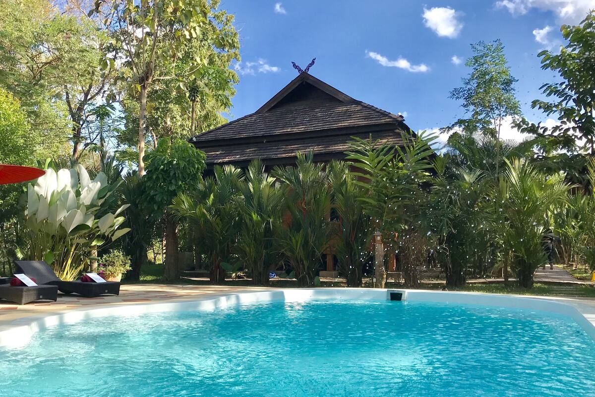 Ein Swimmingpool mit Liegestühlen und Sonnenschirm, umgeben von üppiger Bepflanzung und einem traditionellen Gebäude.