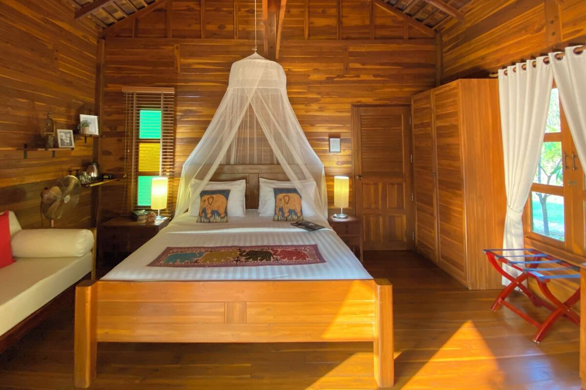 Ein Zimmer aus Holz mit einem Bett, einem Nachttisch, einer Lampe, einem Ventilator und einem Fenster mit Vorhängen.
