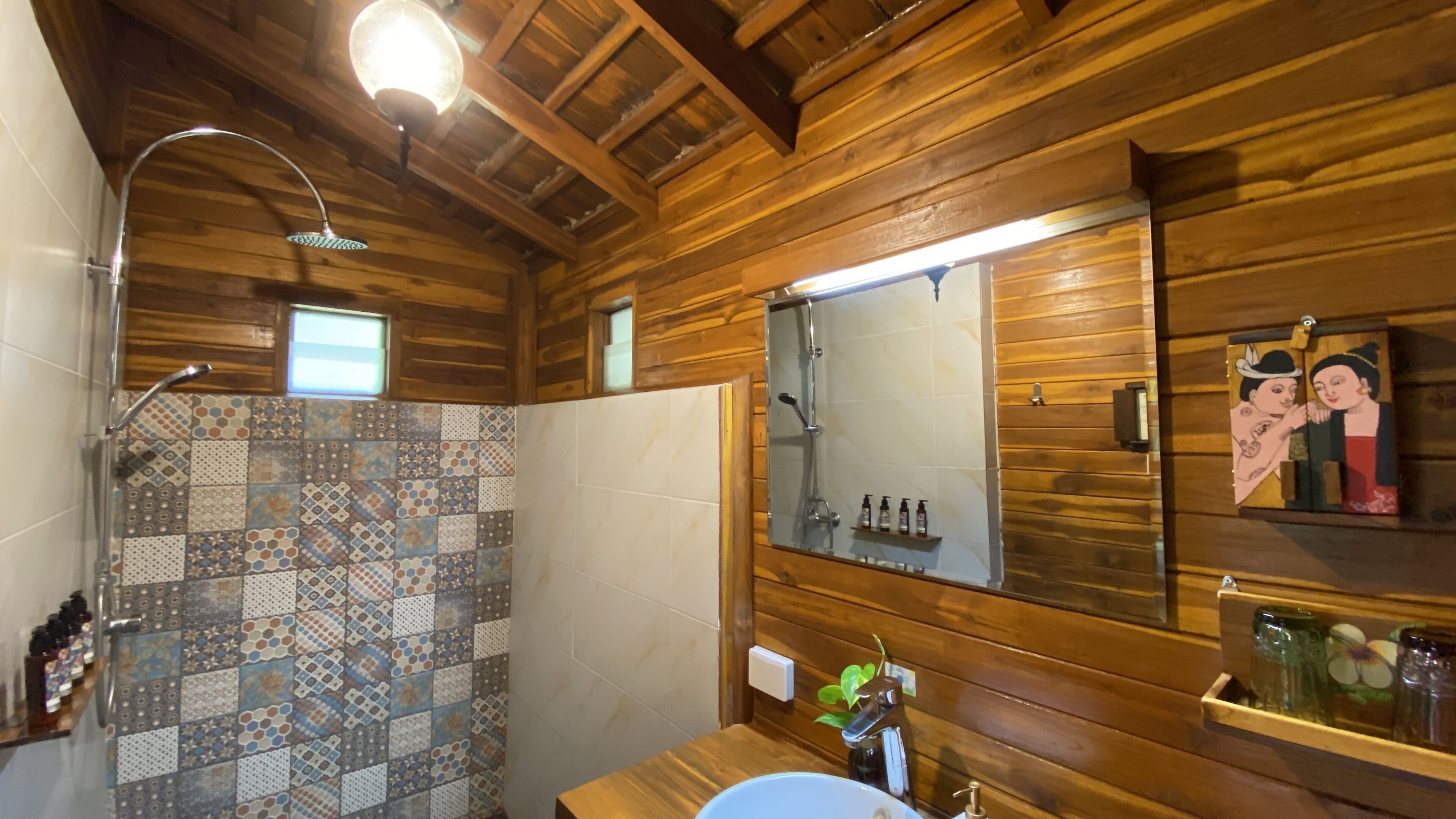 A wooden cabin bathroom with a shower, a sink, a mirror, and a shelf with toiletries.