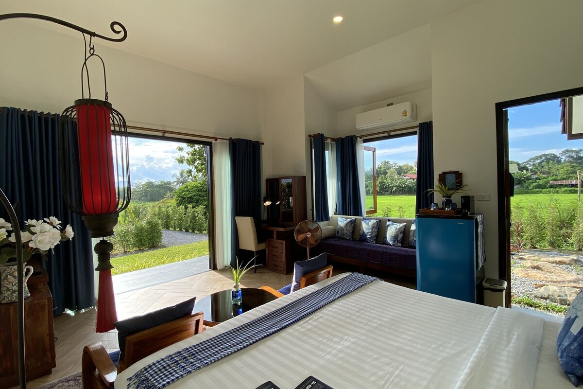 A bedroom with a bed, a desk, a chair, a fan, and a view of the outdoors.