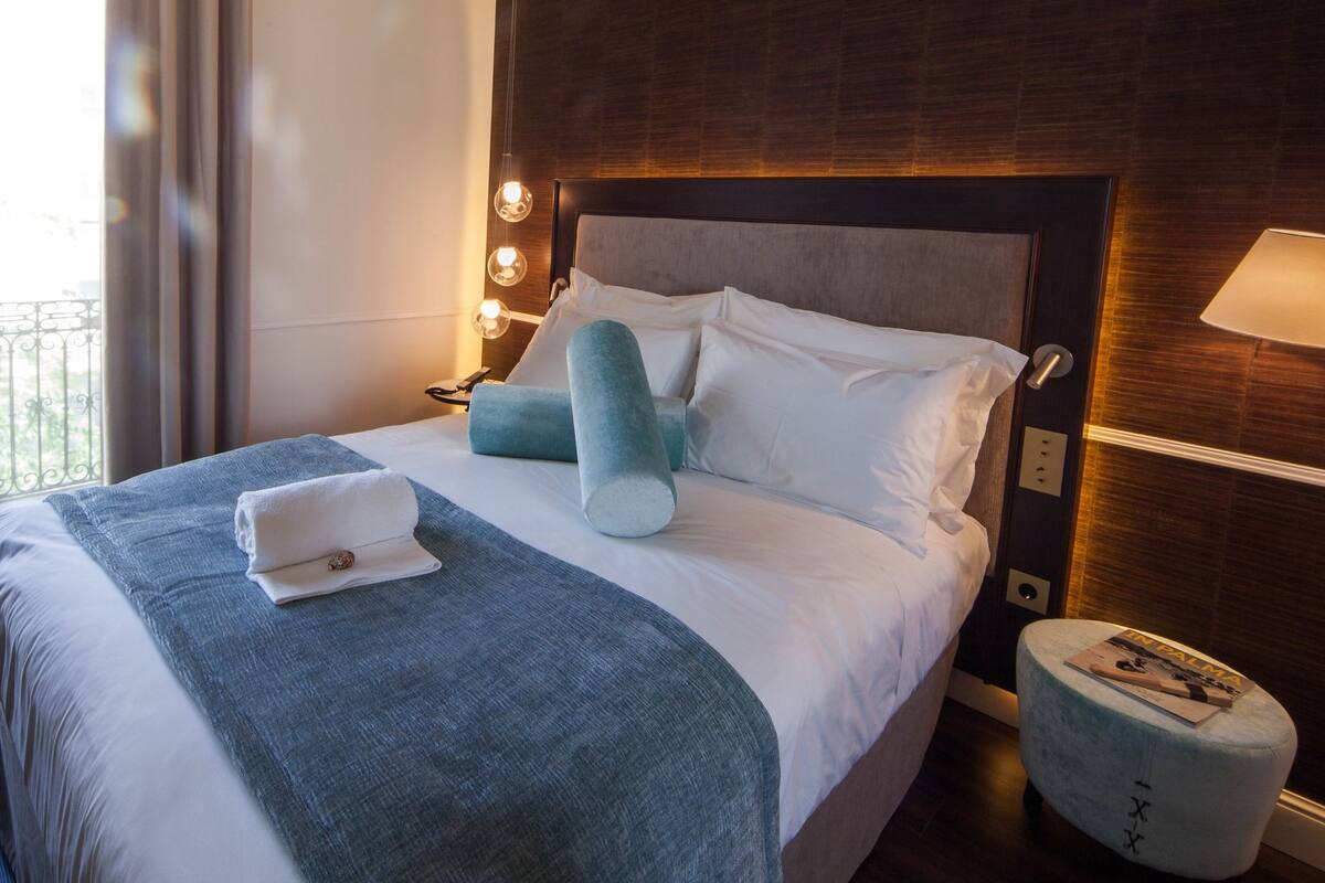 Habitación de hotel con cama, una silla, mesita de noche y vistas al exterior.