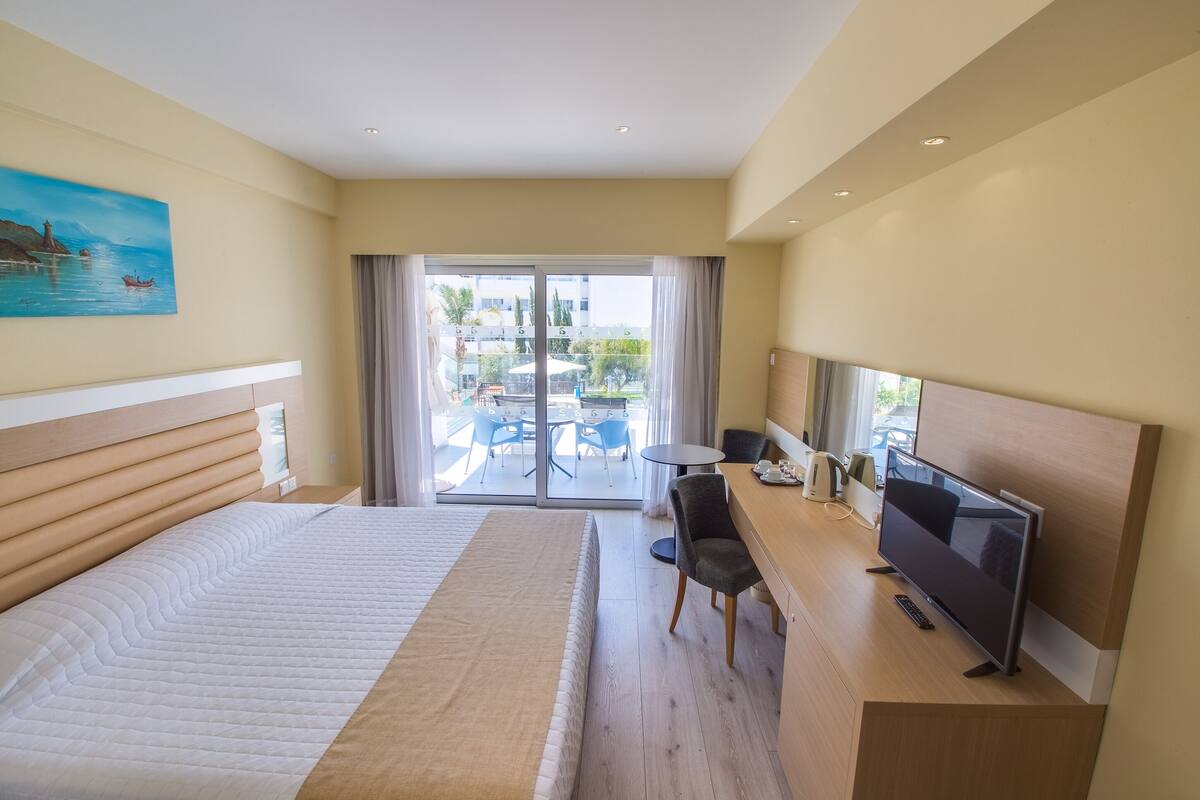 Quarto de hotel com uma cama, uma mesa de trabalho com televisão e varanda com mobiliário de exterior.