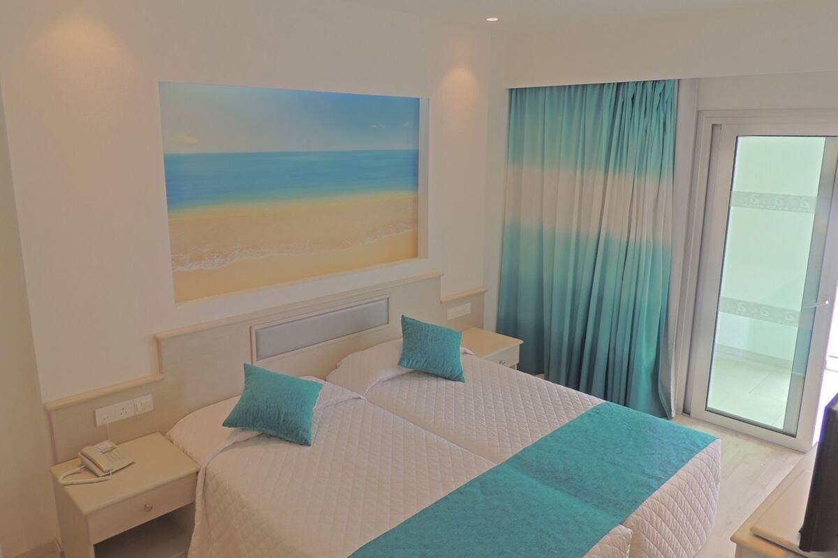 Quarto de hotel com cama, duas almofadas, mesa de cabeceira, pintura de uma praia e janela com cortinas.