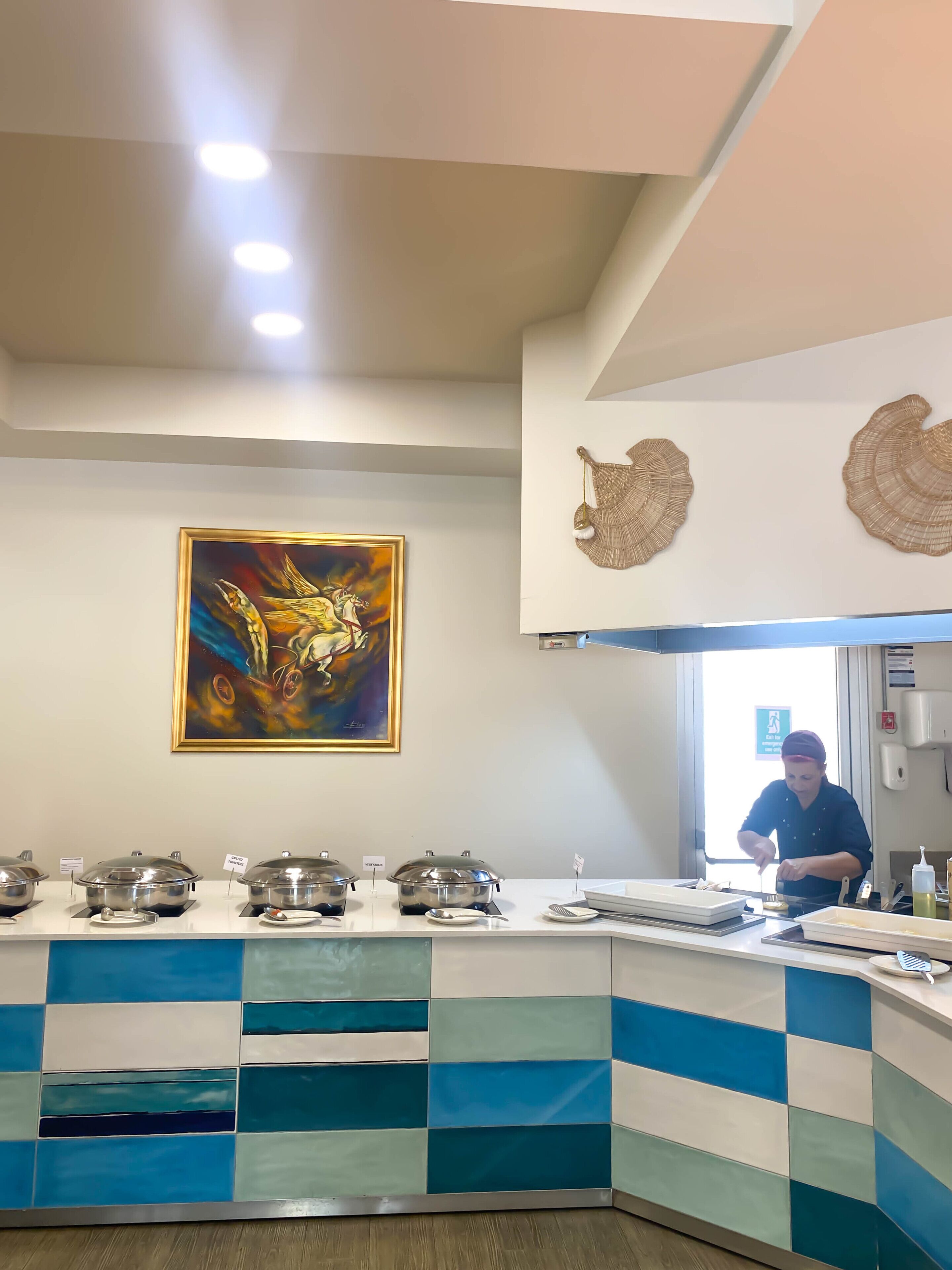 Eine Küchenzeile mit einem Buffet, ein Koch, der Speisen zubereitet, und ein farbenfrohes, abstraktes Gemälde an der Wand.