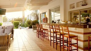 A bar area with a long counter and multiple stools, a ceiling fan, and a view of the outdoors. - Anesis (Ayia Napa)