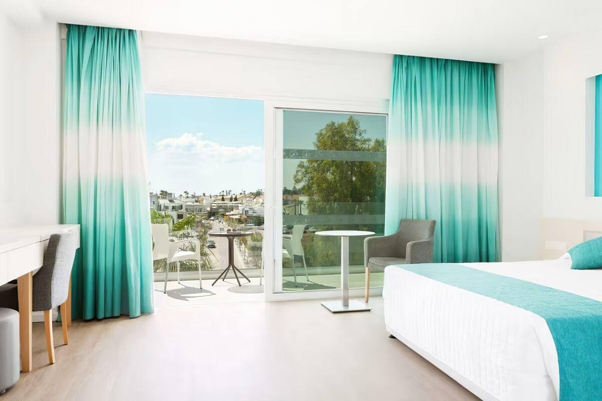 Quarto de hotel com cama, escrivaninha, cadeira, mesa pequena e varanda com vista para a cidade.