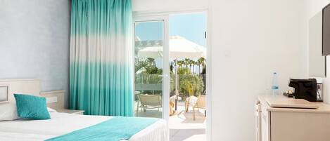 A hotel room with a bed, a desk, a chair, and a view of the outdoor area including a pool and umbrellas.