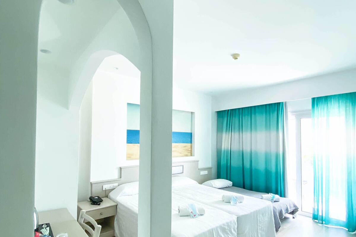 Quarto de hotel com duas camas, uma escrivaninha e uma janela grande com cortinas.