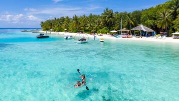 Una playa tropical con agua turquesa cristalina, una pareja haciendo kayak y un resort frente a la playa con sombrillas y sillas de descanso.