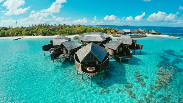 Wasserbungalows über türkisfarbenem Wasser, mit einem Strand im Hintergrund.