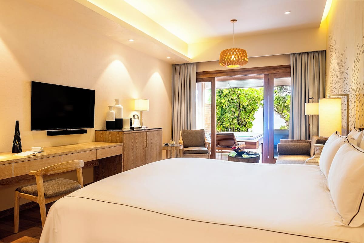A hotel room with a large bed, a desk with a TV, a chair, and a sofa. There is a view of greenery outside the window.