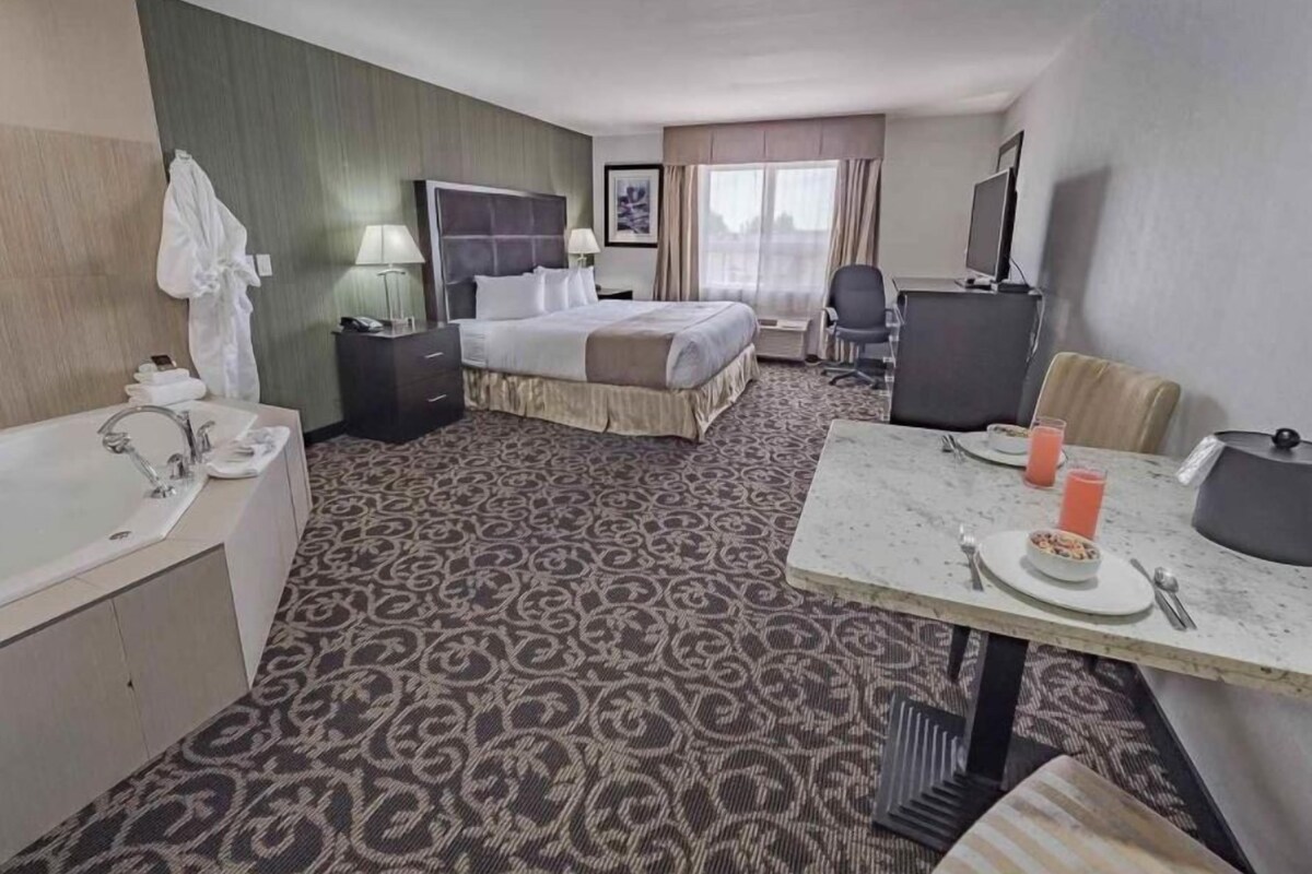 Quarto de hotel com cama, escrivaninha com cadeira, mesinha com prato e copos, pia e espelho.