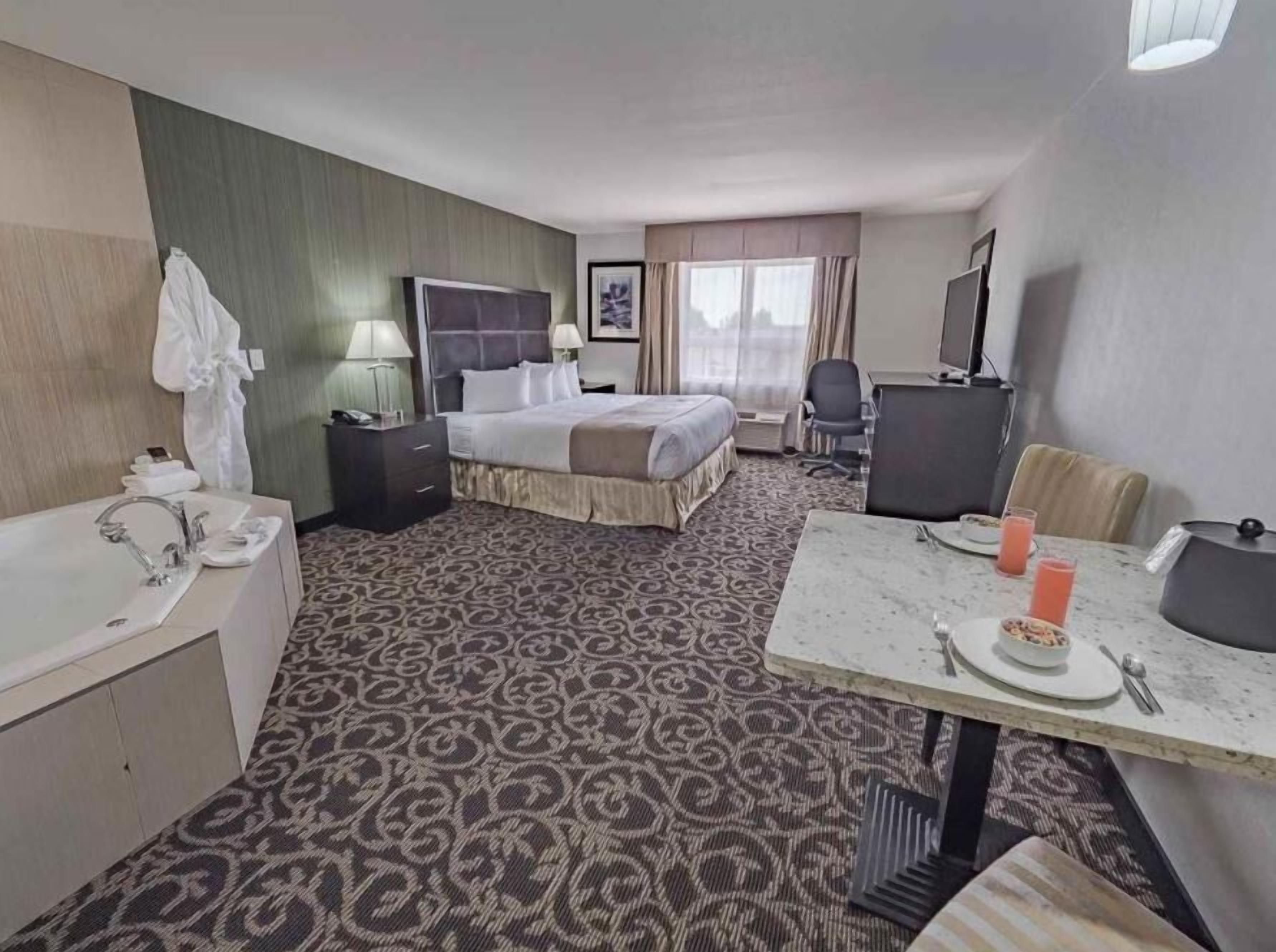 Quarto de hotel com cama, escrivaninha com cadeira, mesinha com prato e copos, pia e espelho.