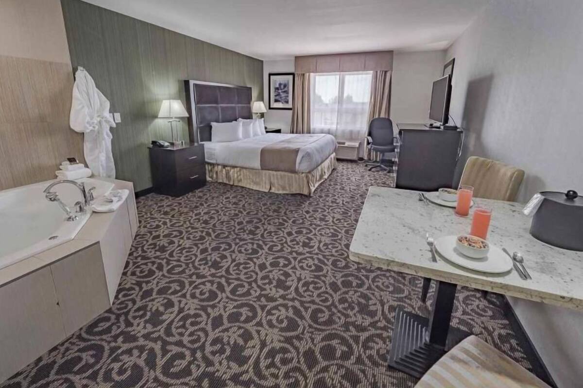 Quarto de hotel com cama, escrivaninha com cadeira, mesinha com prato e copos, pia e espelho.