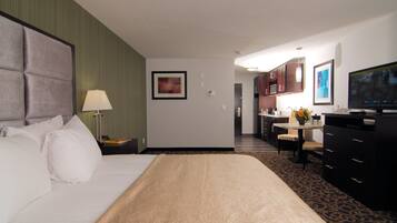 A hotel room with a large bed, a desk, a chair, a TV, and a kitchen area.