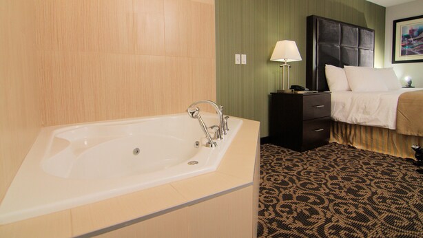 A hotel room with a large bathtub, a bed with a headboard, a bedside table with a lamp, and a patterned carpet. - Paradise Inn & Kitchenette Suites (Redwater)