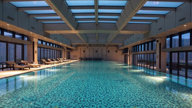Une piscine intérieure dotée d’un toit en verre, de chaises longues disposées le long des bords et de grandes fenêtres.
