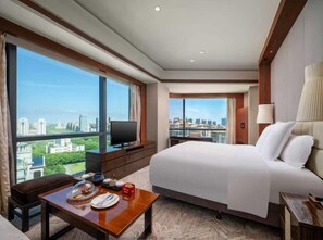 A hotel room with a large bed, a TV on a wooden cabinet, a dining table with a plate and a glass, and a view of a cityscape through large windows.