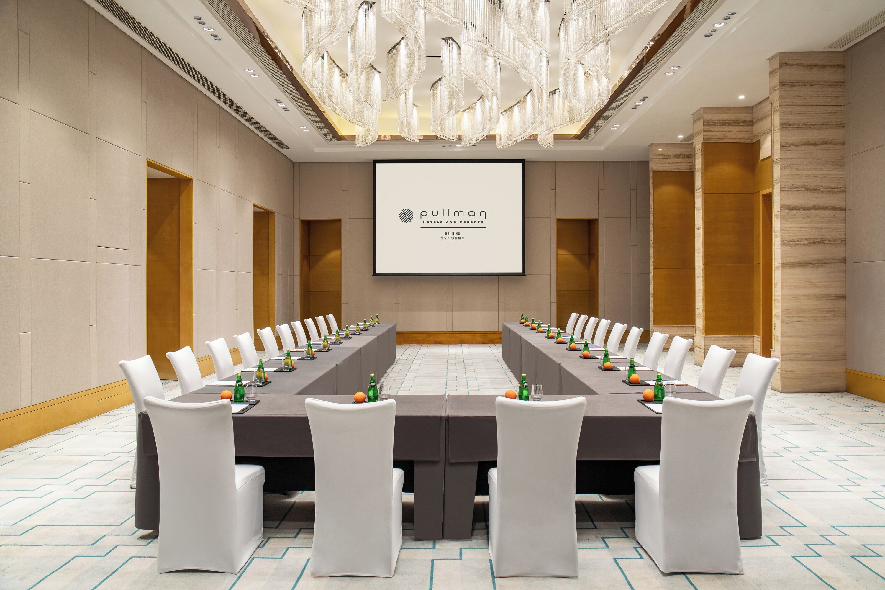 A conference room with a long table, white chairs, and a screen displaying the hotel name.