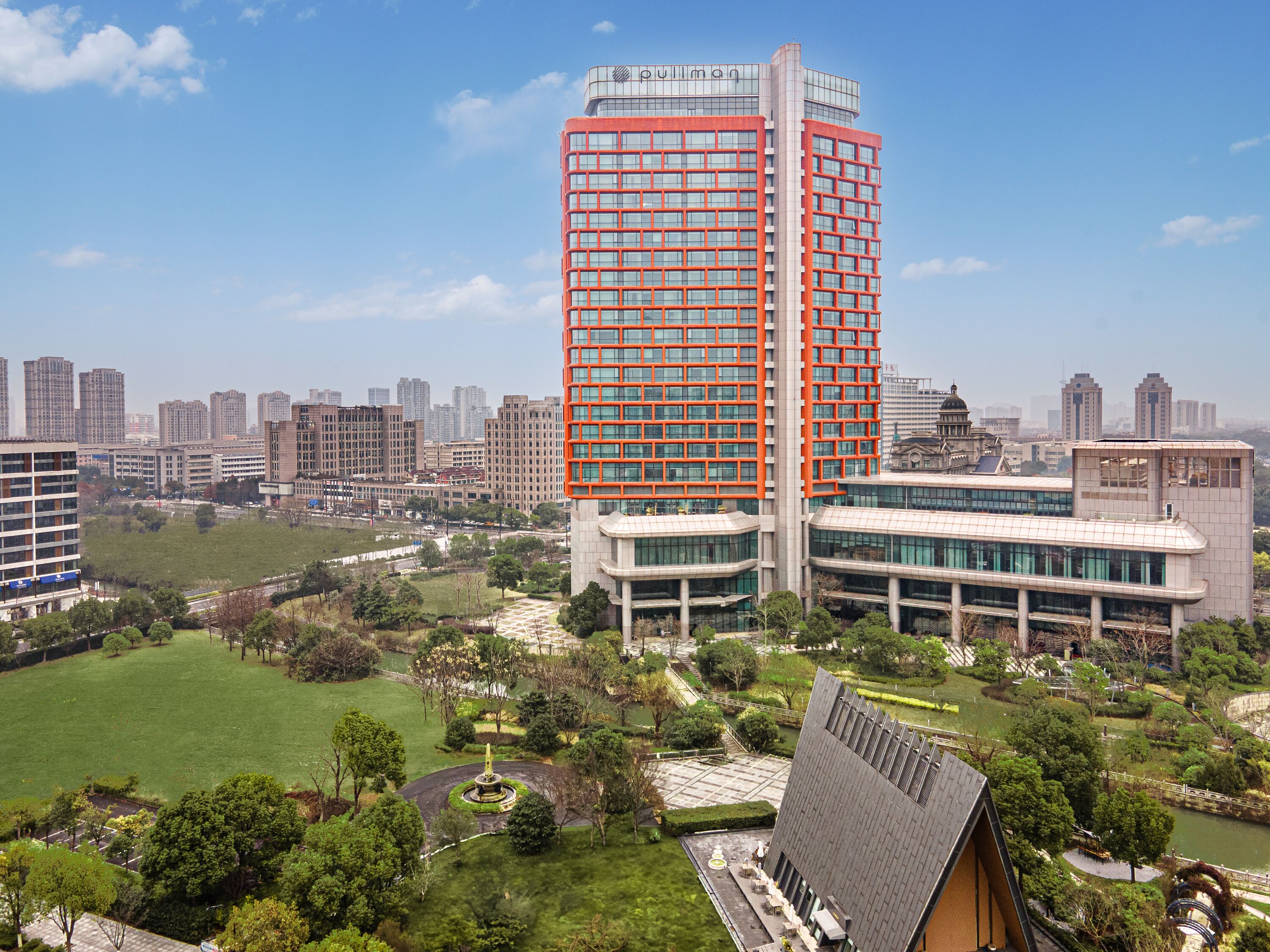 A modern high-rise building with a prominent red facade, surrounded by a well-maintained park with a central fountain and pathways.