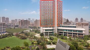 A modern high-rise building with a prominent red facade, surrounded by a well-maintained park with a central fountain and pathways.