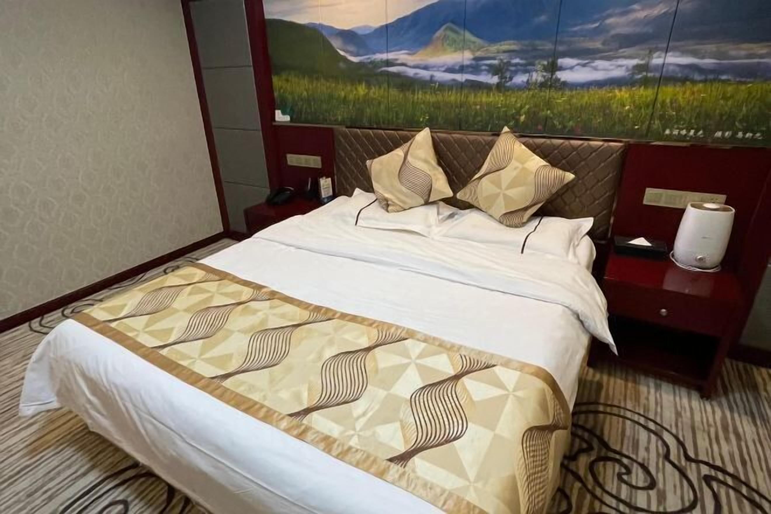 A hotel room with a large bed, a headboard with a landscape painting, a red nightstand with a lamp, and a patterned carpet.