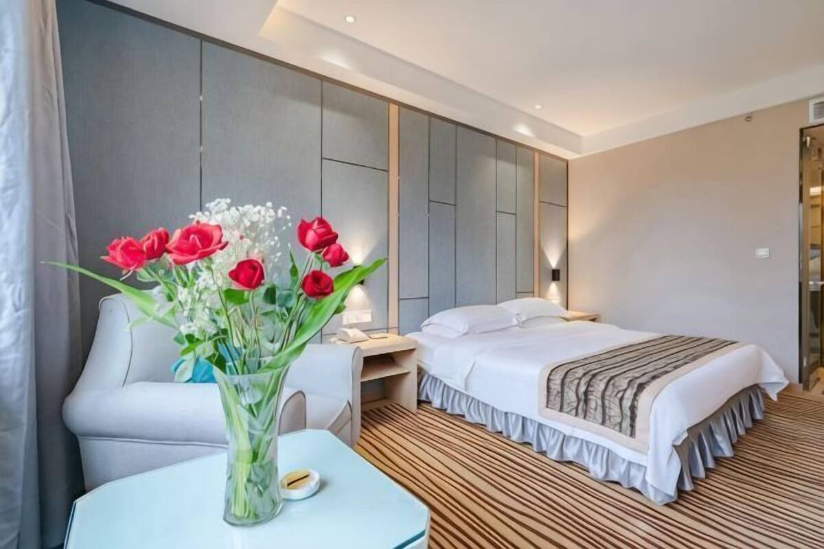 A hotel room with a bed, bedside table, chair, and a vase with flowers.