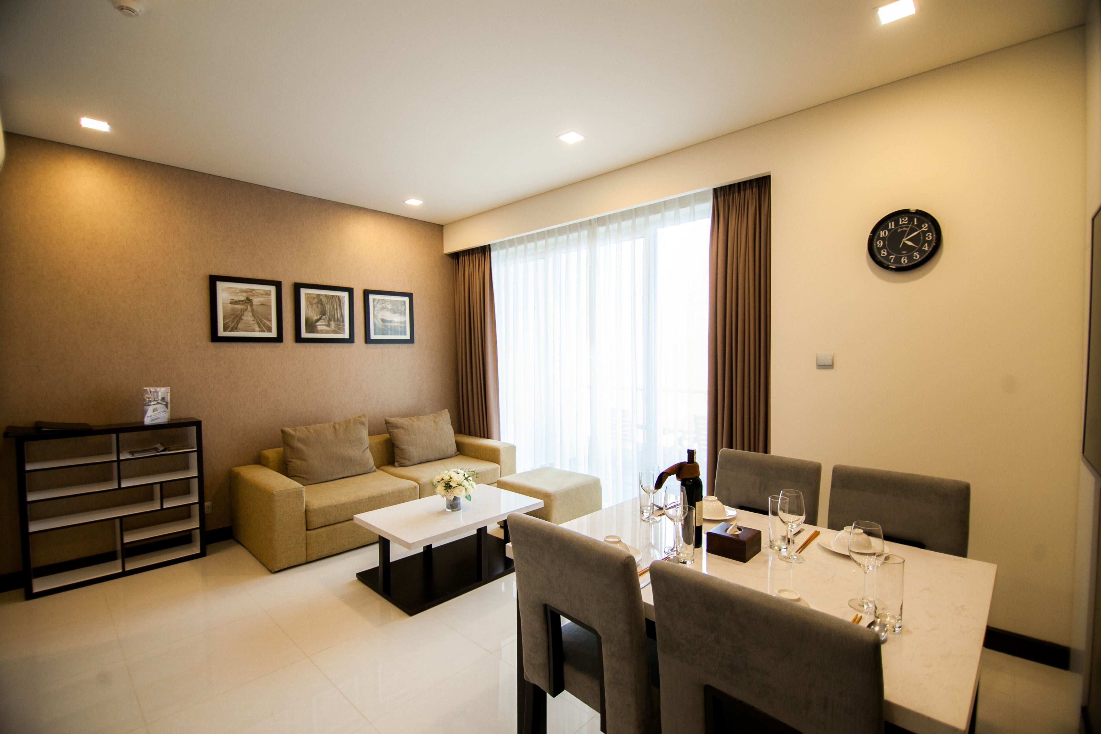 A modern living room with a sofa, coffee table, and dining table set for four. There are three framed pictures on the wall, a clock, and a window with curtains.