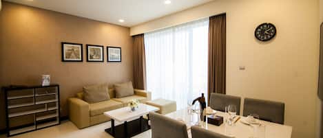 A modern living room with a sofa, coffee table, and dining table set for four. There are three framed pictures on the wall, a clock, and a window with curtains.