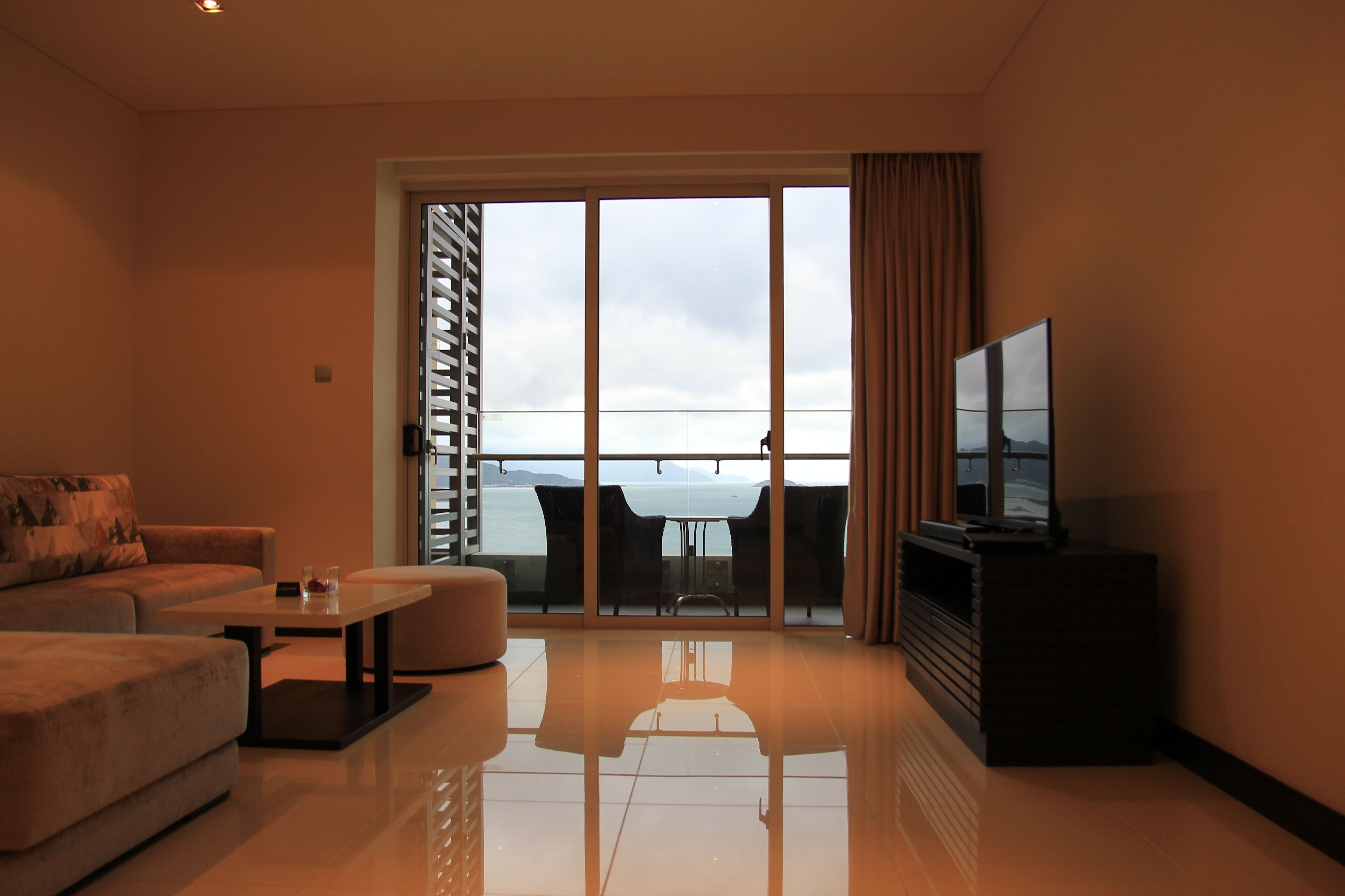 A modern living room with a sofa, a coffee table, a flat-screen TV, and a balcony with a view of the sea.