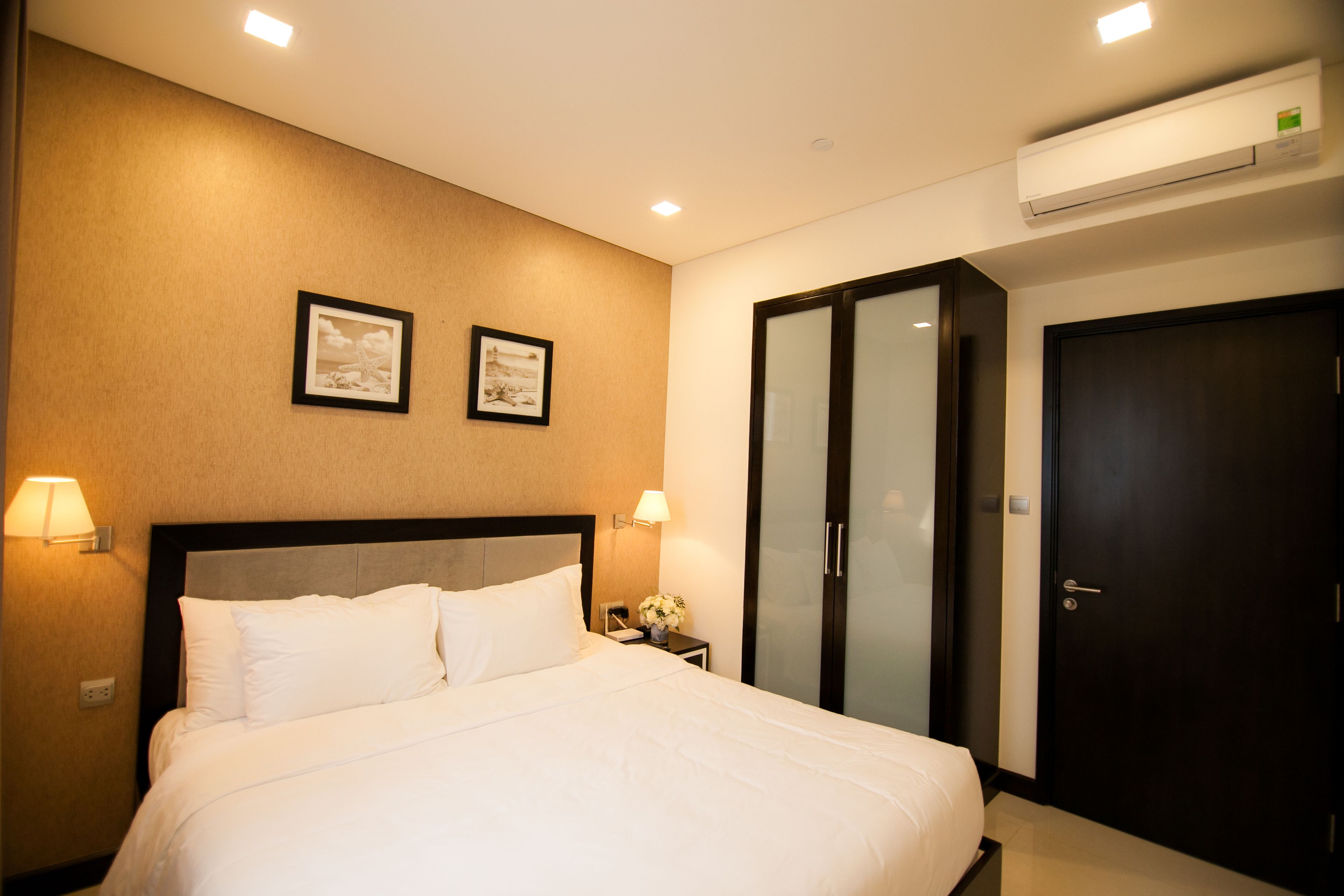 A hotel room with a large bed, two wall-mounted lamps, a nightstand with a vase of flowers, a wardrobe, and an air conditioning unit.