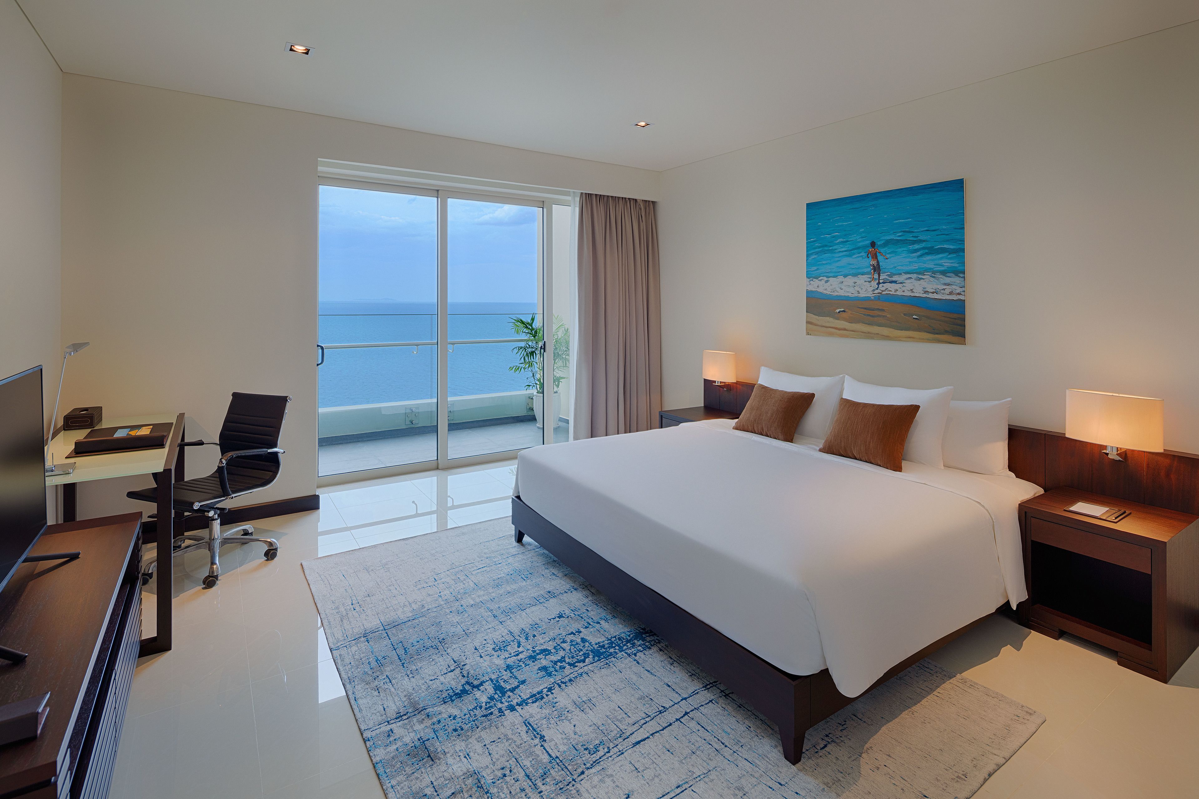 A modern hotel room with a large bed, a desk with a chair, a TV, and a view of the sea through a sliding door.