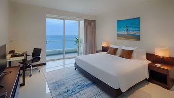 A modern hotel room with a large bed, a desk with a chair, a TV, and a view of the sea through a sliding door.