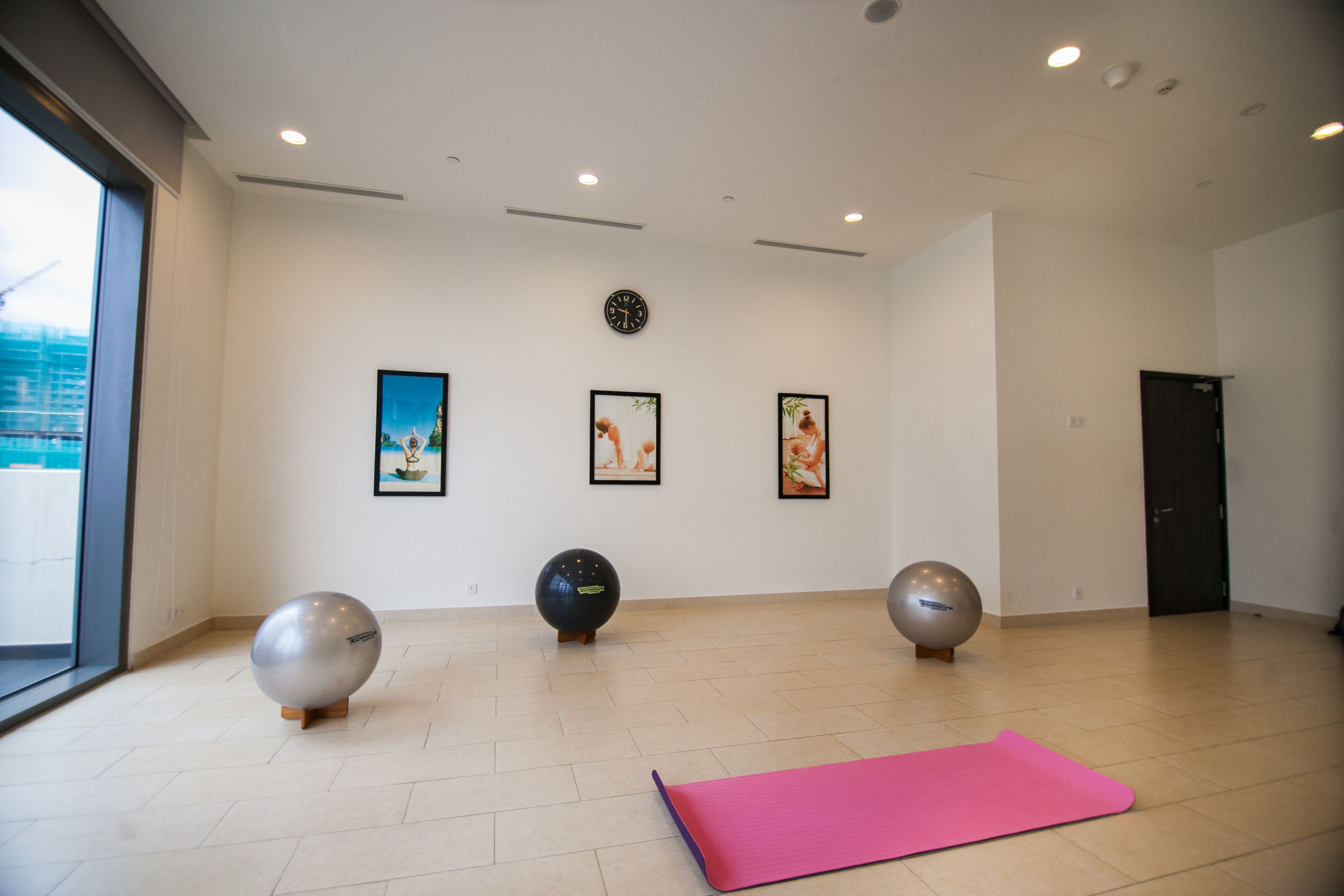 A spacious room with a large window, a pink exercise mat, and three exercise balls against the wall.